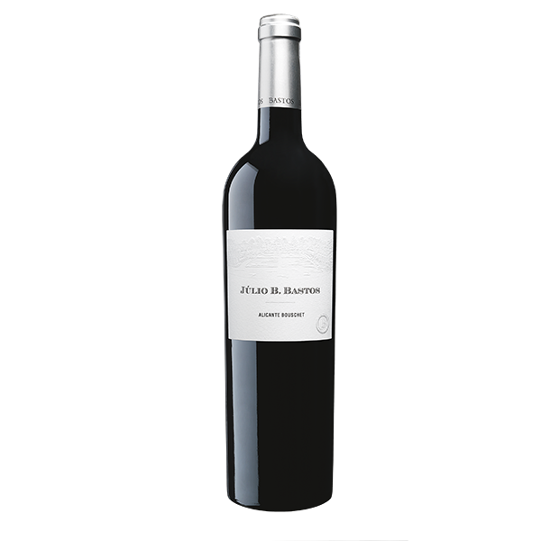 Taça de vinho tinto Julio Bastos Alicante Bouschet da marca Dona Maria com garrafa ao fundo.